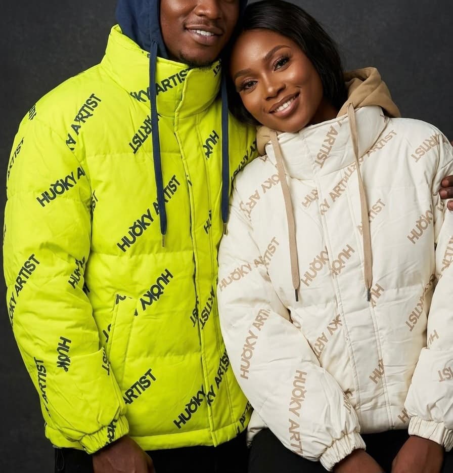 Man and woman wearing unisex all-over graphic puffer jackets with high collars and statement typography